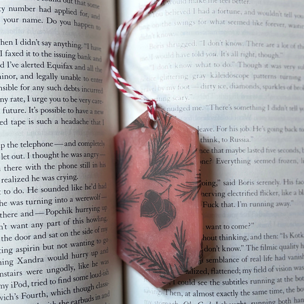 Decorative pink wax bookmark with christmas tree leaves and acorns drawn on it, resting on an open book