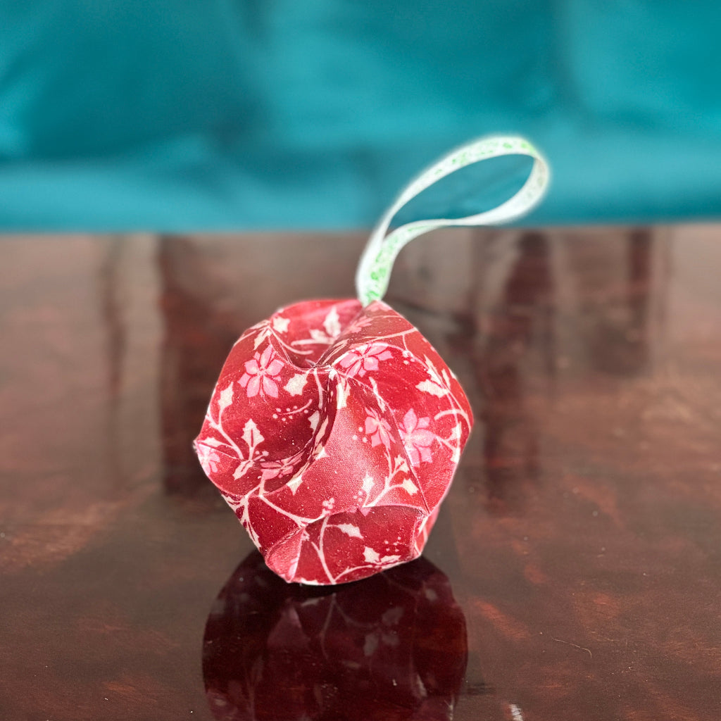 Red floral waxed fabric Christmas bauble ornament with a green string on a reflective surface.